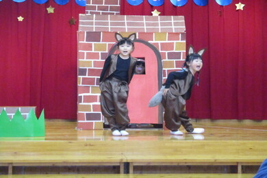 茶色い衣装を着て猫耳をつけた二人の園児がステージの上のレンガの家の手前でポーズをとっている写真