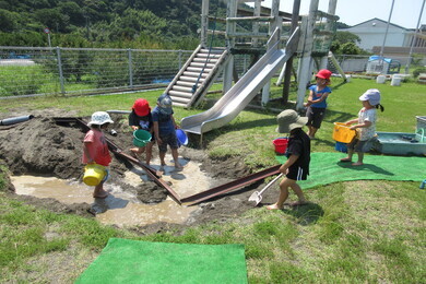 屋外の芝生の生えた公園で滑り台の手前に大きな穴があり、その中や周りに園児たちがいる写真