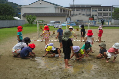 屋外で泥の中を園児たちが裸足で屈みながら遊んでいる写真