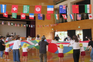 世界中の国旗が体育館の上部からかけられており、カラフルな大きな一枚の布を大勢の園児たちと大人たちが一緒に持っている写真