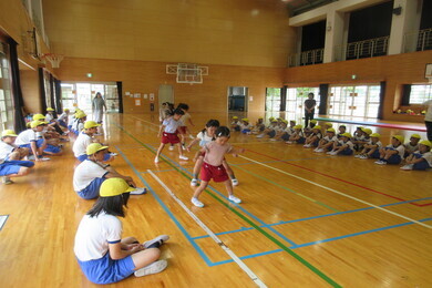 反復横跳びに挑戦している園児たちとその様子を座って見ている小学生たちの写真