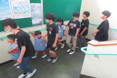 幼児たちが小学校で6年生に手を繋がれて避難経路を歩いている写真