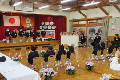 制服を着た園児たちがステージの上と体育館の広場に置かれた椅子の上に座っている写真