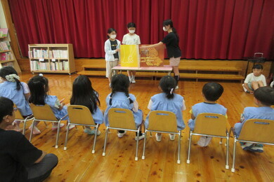 園児たちが椅子に座りながら小学生の絵本の読み聞かせを聞いている後ろからの写真