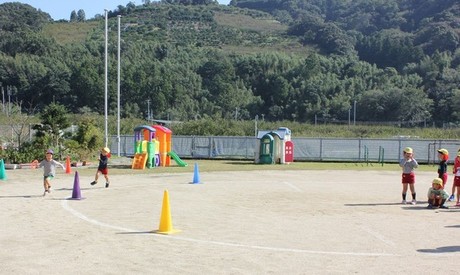 園児たちが広い園庭で運動会ごっこやリレー競走をして楽しそうに遊んでいる写真