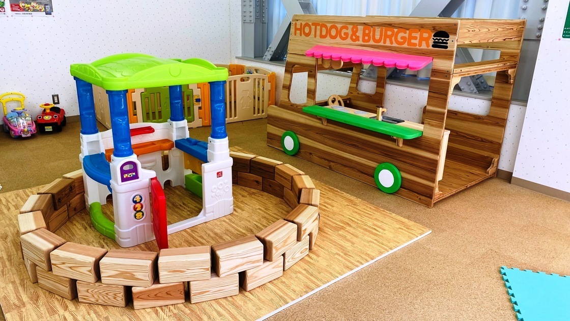 コルクタイルを張った床の上にジョイントマットが敷かれており、その上にプラスチックや木でできた遊具が置かれている写真