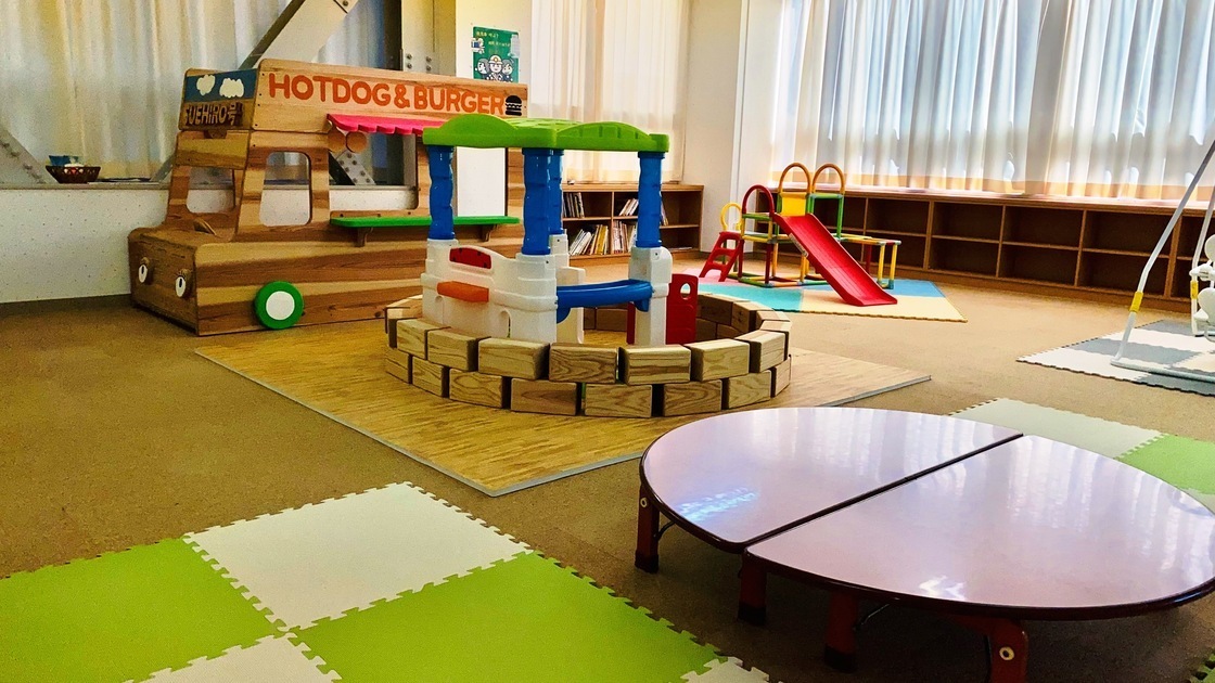 コルクタイルを張った床の上にジョイントマットが敷かれており、その上にプラスチックや木でできた遊具とテーブルが置かれている写真