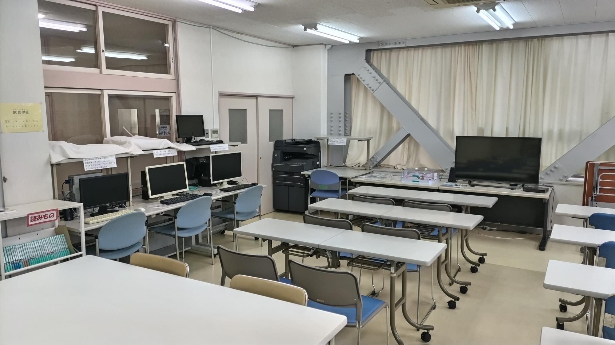 クッションフロアの床の上に長机と椅子が並べられており、その他にパソコンデスクとその上にパソコン、テーブルとその上にテレビが置かれている写真