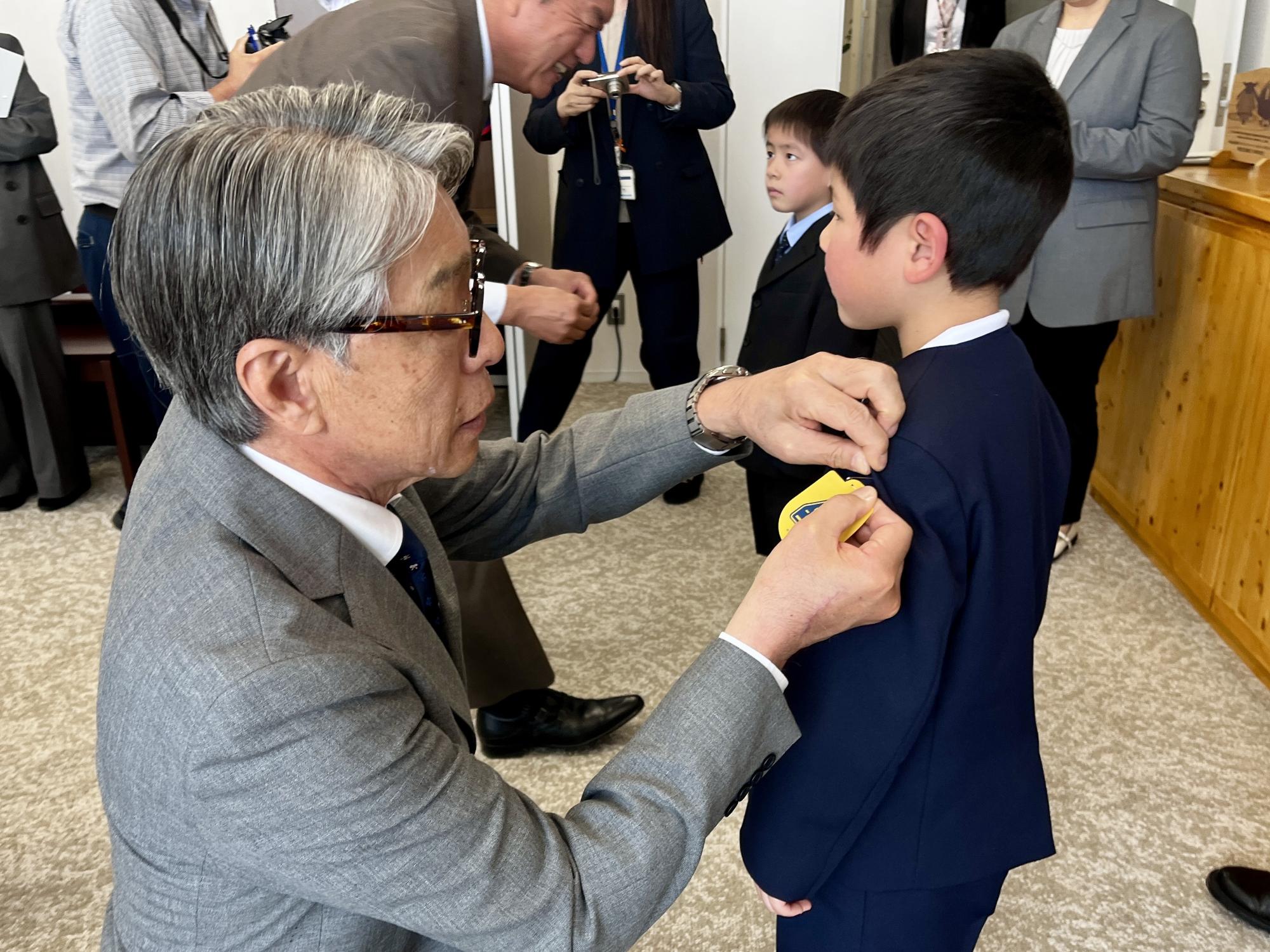 スーツ姿の男性が、目の前に立った男の子の袖に、黄色いワッペンを取り付けてあげている写真