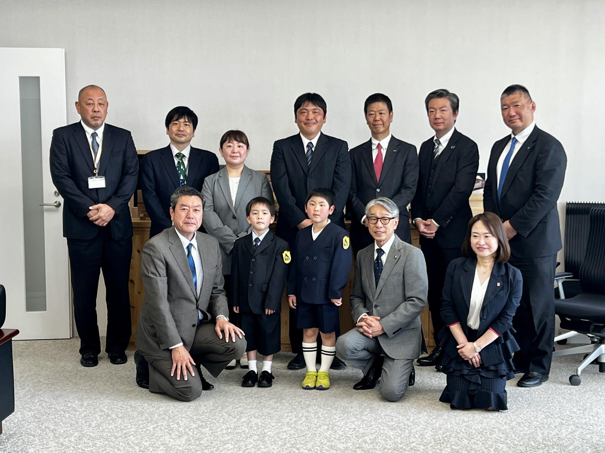 黄色いワッペンを袖につけた小学生たちを囲むようにスーツ姿の男女が数列に分かれて整列し、全員で正面を向いている写真