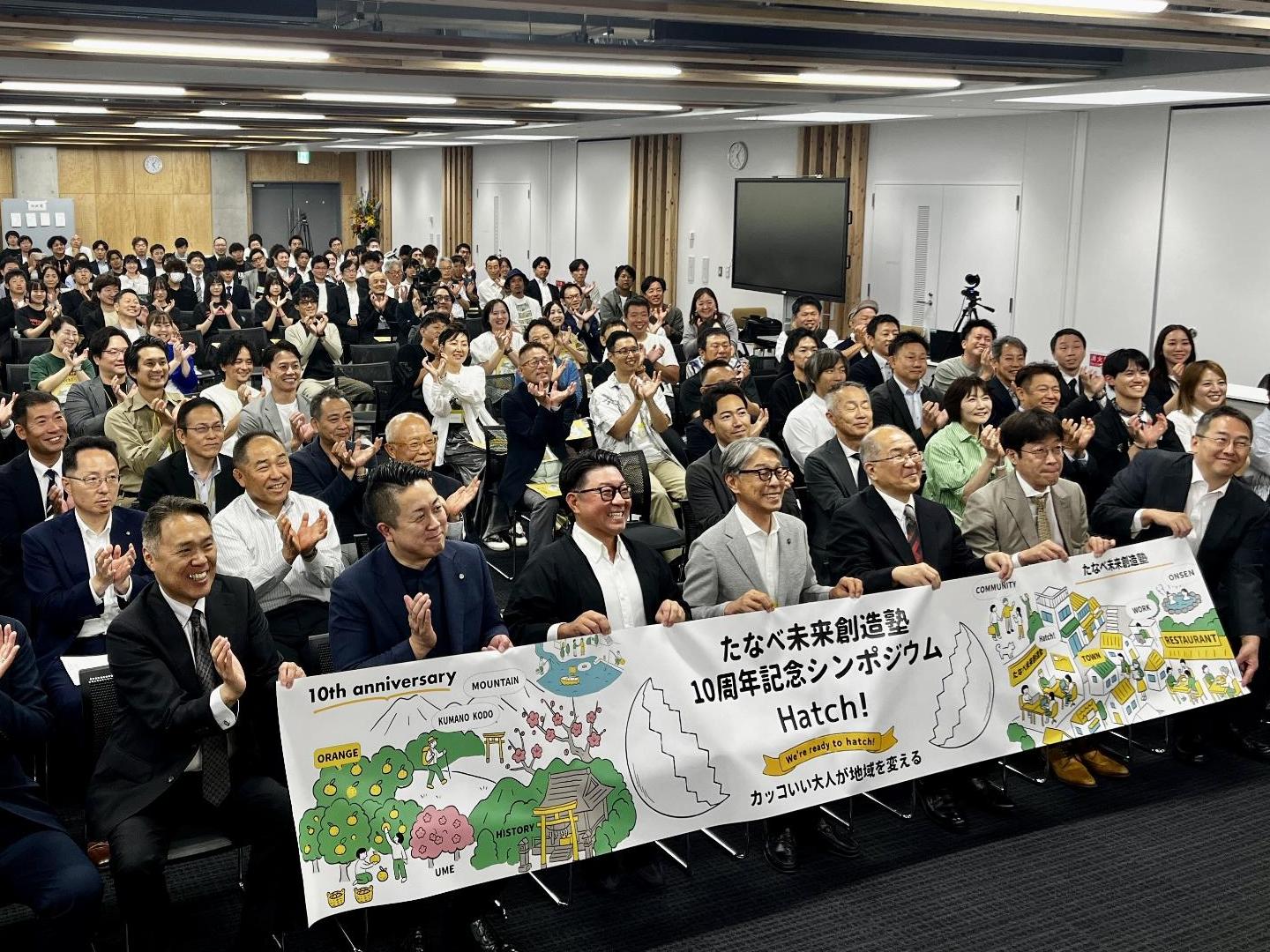 200人ほどの観客が室内で正面を向いて着席し、最前列の人々がタペストリーを持ち合い記念撮影をしている写真