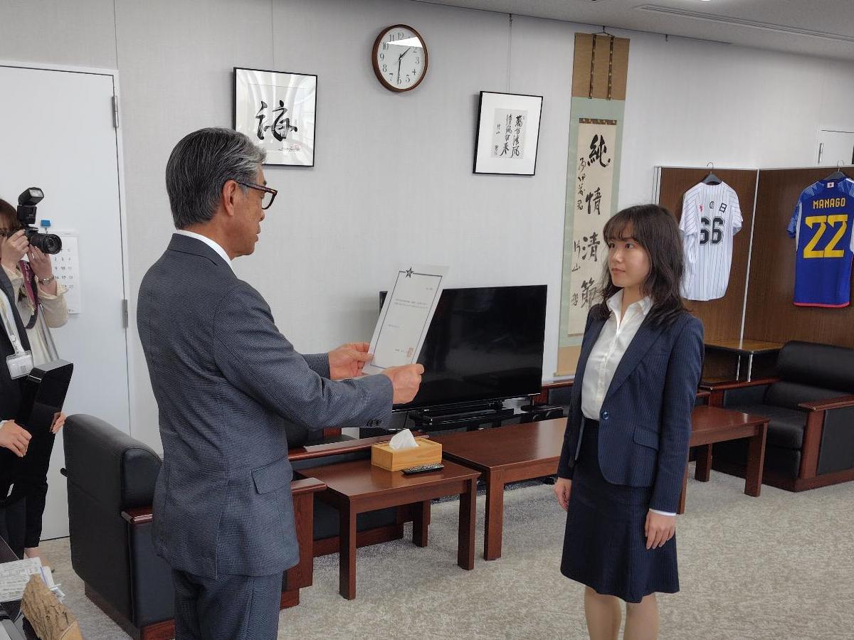 スーツ姿の女性が、前方に立つ人物から書類を両手で受け取っている写真