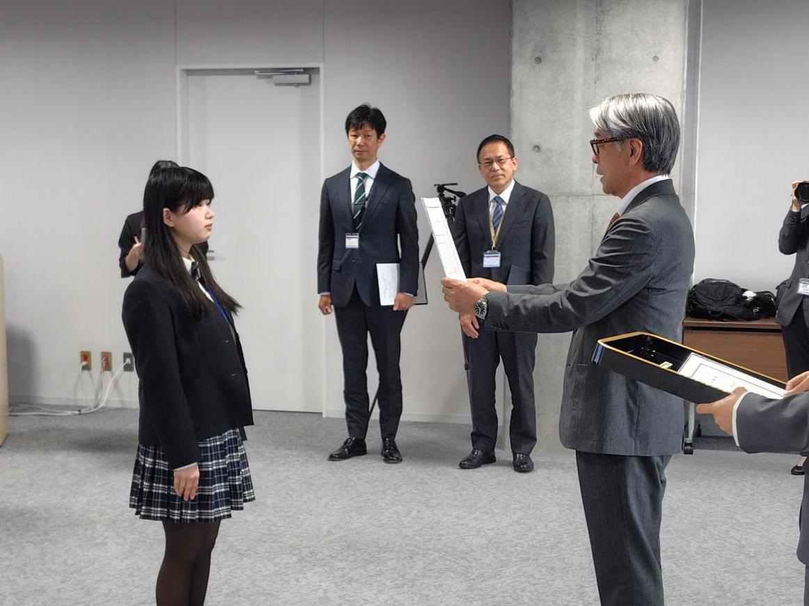 制服姿の女性とスーツ姿の男性が並んでたち、委嘱状らしきものを手渡されている写真