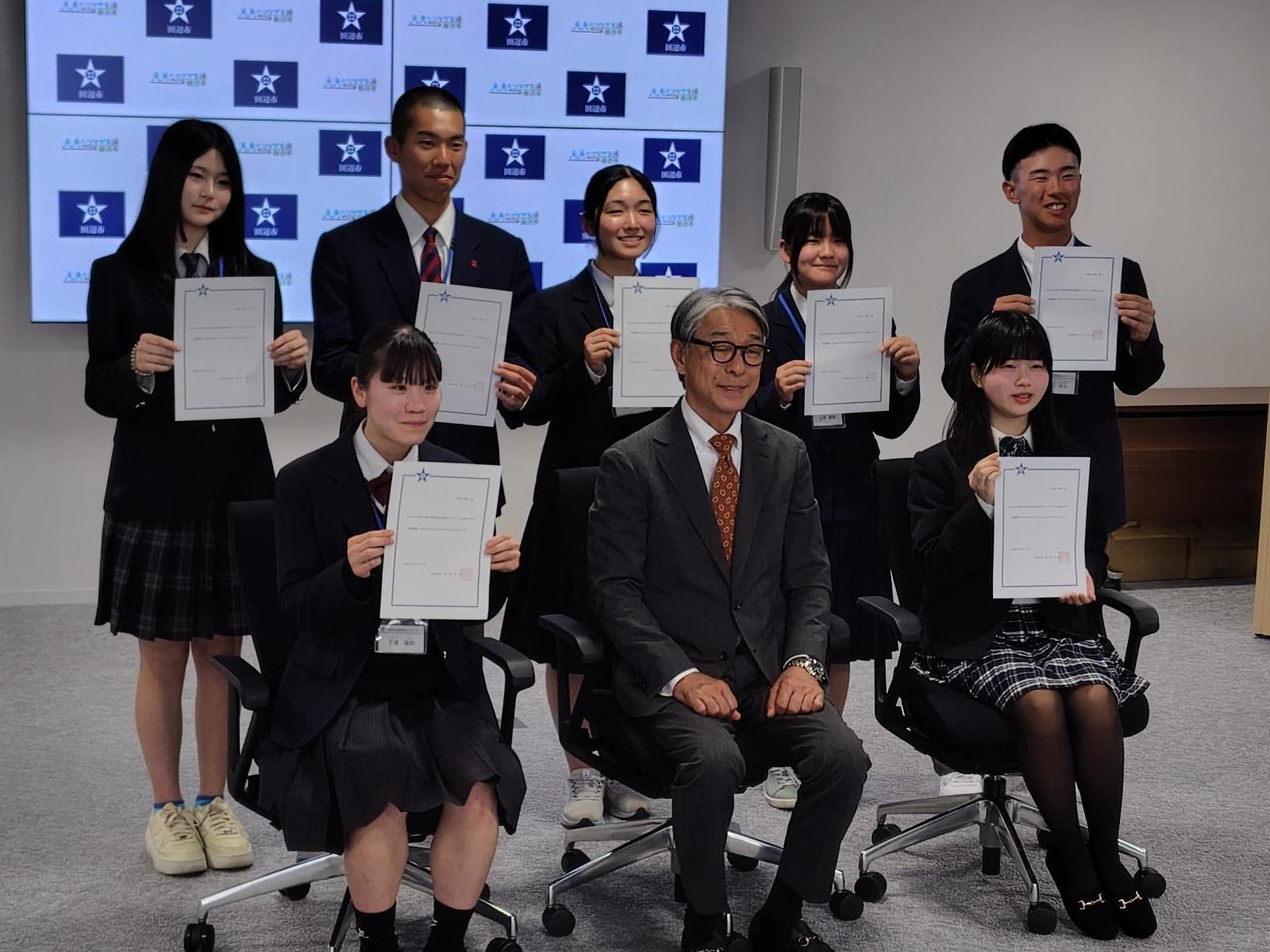 前方の椅子にスーツ姿の男性が座り、その後ろに高校生たちが数列に分かれて整列し、全員で正面を向いている写真