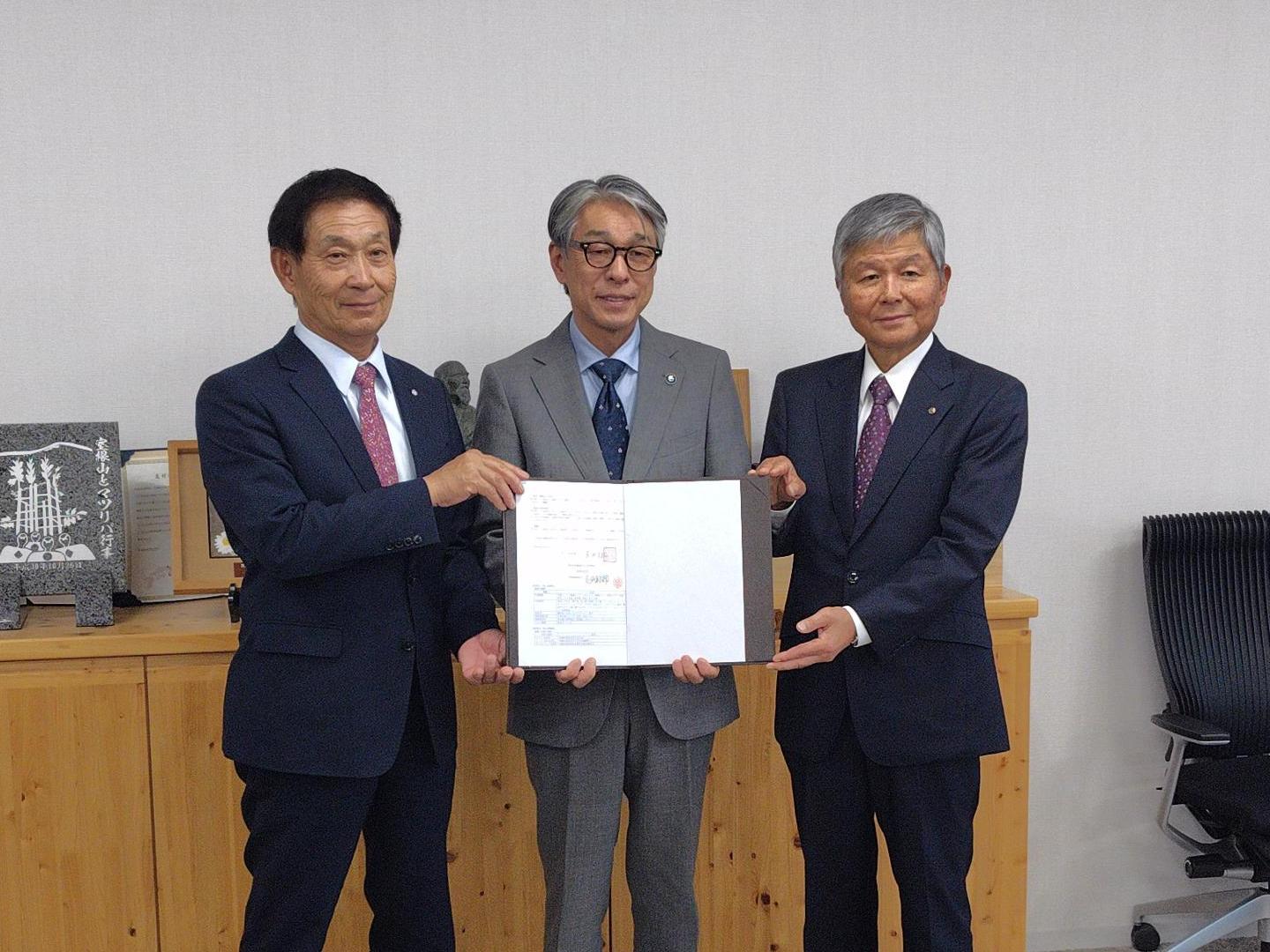 スーツ姿の男性たちが立ち、協定書を持ち合って正面を向いている写真