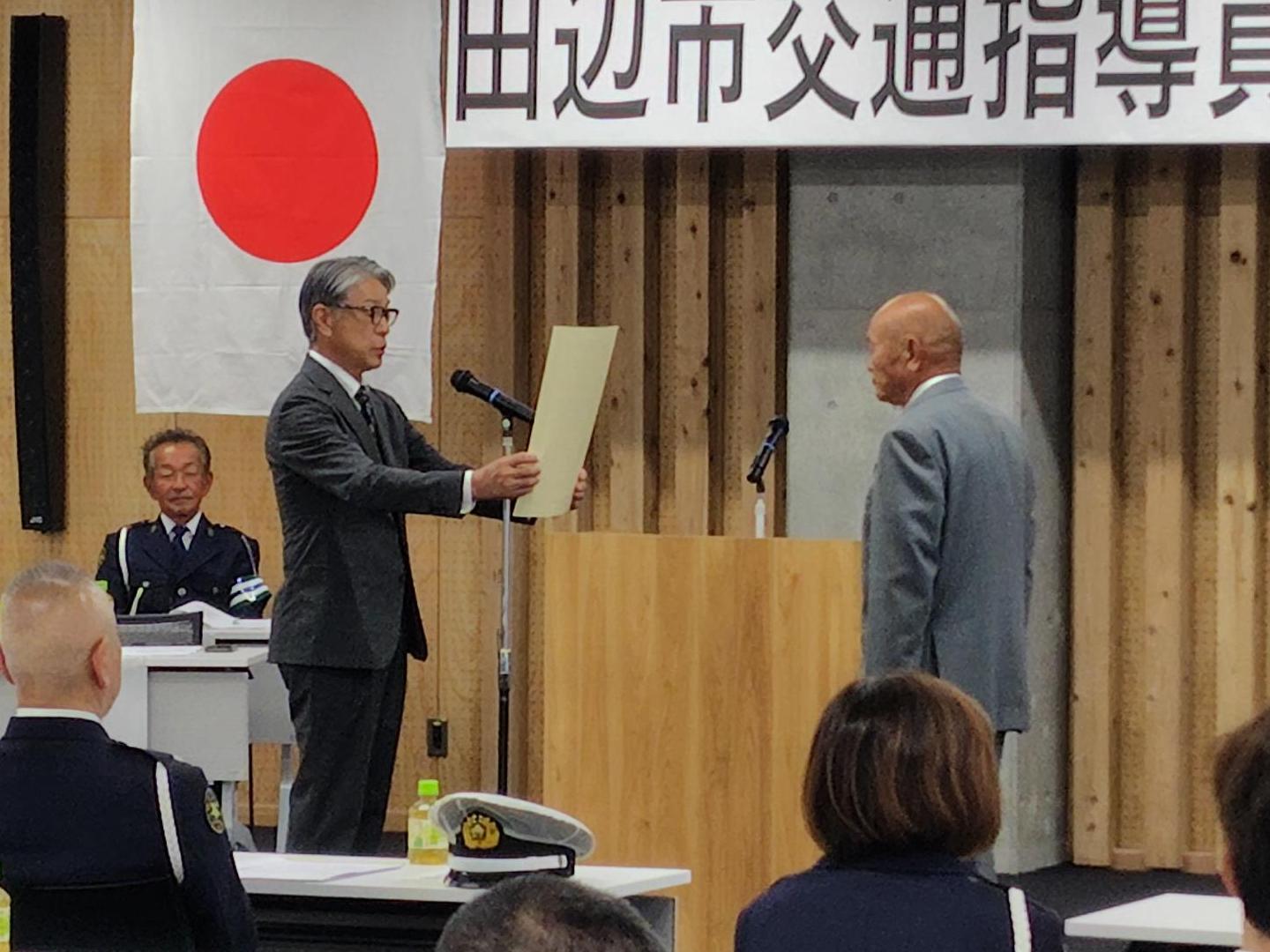 会場にて、前方の台にスーツ姿の男性が立ち、代表者から賞状を授与されている写真