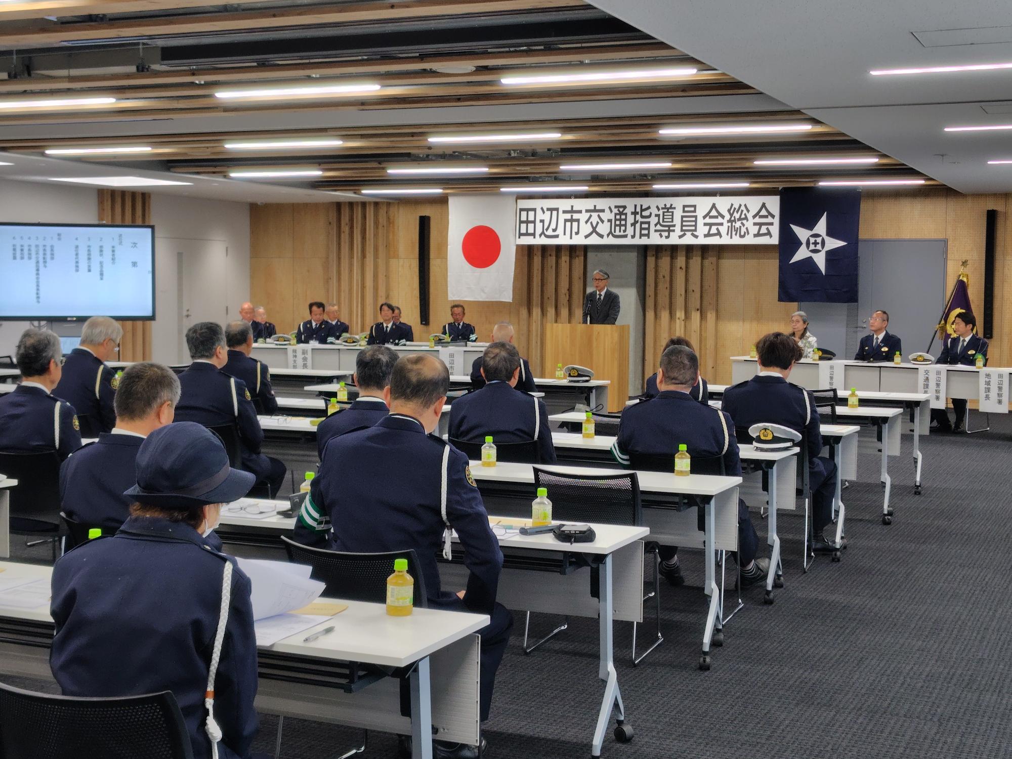 前方にスーツ姿の男性が立っており、並べられた机に座って多くの出席者が話を聞いている写真