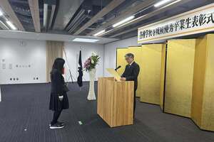 表彰式の会場にて、マイクの前に立つ男性から表彰状を受け取っている写真