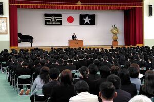 体育館の卒業式にて、演台の前に立つ男性の前に大勢の人が座っている写真