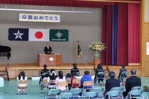 男性が壇上にたち、卒業生や参加者が前の椅子に座っている写真