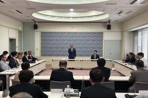 会議室にて、スーツを着た人たちがコの字型に並んだテーブルに着席し、会議をしている写真