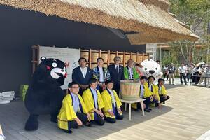 緑の法被を着た男性、関係者、そして2体の着ぐるみキャラクターが、漬物桶を囲んで2列に並んでいる写真