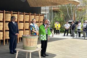 緑色の法被を着た男性が漬物桶の横でマイクをもって話をし、周りで人々が聞いている写真