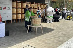 緑の法被を着た男性が大きな漬物桶で作業をしており、周囲を2体の着ぐるみキャラクターと人々がその様子を見ている写真