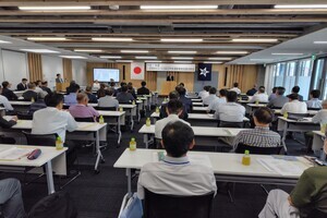 会議室で人が、並べられた机と椅子があり、座って話を聞いている写真