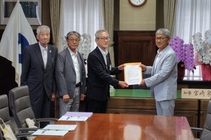 知事室にて、左側にスーツを着た3人の男性と右側に立つグレーのスーツの男性が両手で1枚の紙を持って正面に提示している写真