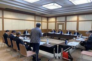 会議室でロの字型の机が配置されており、出席者が着席している中でスーツ姿の男性が立ち上がっている写真