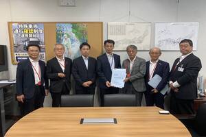 スーツ姿の男性たちが並んで立っており、その中央にいる2人の男性が書類を一緒に手に持って正面を向いている写真