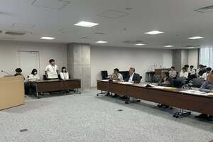 会場の前方白い服を着た男性が立っており、その横の座席に座った人々が注目している写真