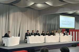 会場の前方に設置された長い机に、男性が横一列に並んで座っている写真