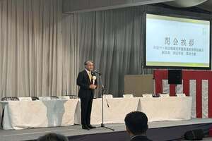 スーツ姿の男性がマイクの前に立ち、来賓挨拶や講演などのスピーチを行っている様子の写真