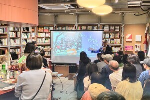 壁一面に本棚がある室内で前方モニター画面を使って説明を行っている人物と、その前で話を聞いている写真