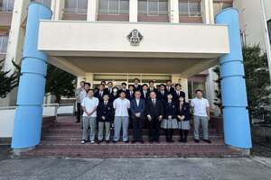 校門前にて、制服姿の生徒たちと数名の大人たちが、前後の列に分かれて並んでいる写真