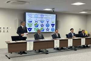 会議室で前方の机にスーツ姿の男性たちが並んで座り、そのうちの一人が資料を説明している写真