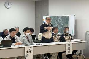 長いテーブルの前に立ちマイクを待って話している男性の写真