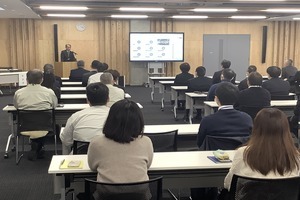 会議室の左前方に人がたっており、着席した大勢の参加者がその話を座って聴いている写真