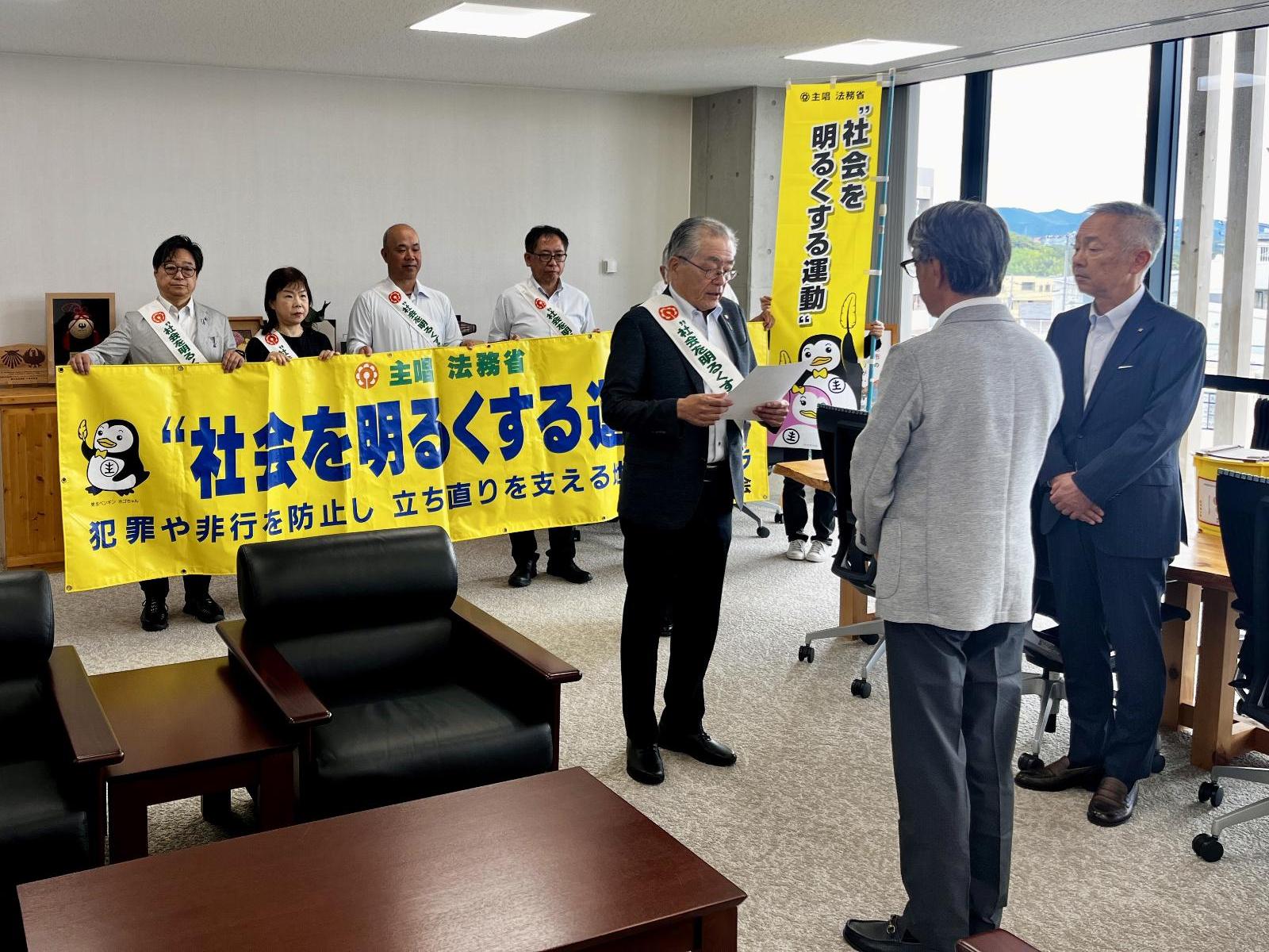 会議室の前方で黄色の横断幕を男女数名が掲げ、その前でスーツの男性3名が向かい合い、うち正面を向いている男性が紙を見てメッセージを読み上げている写真