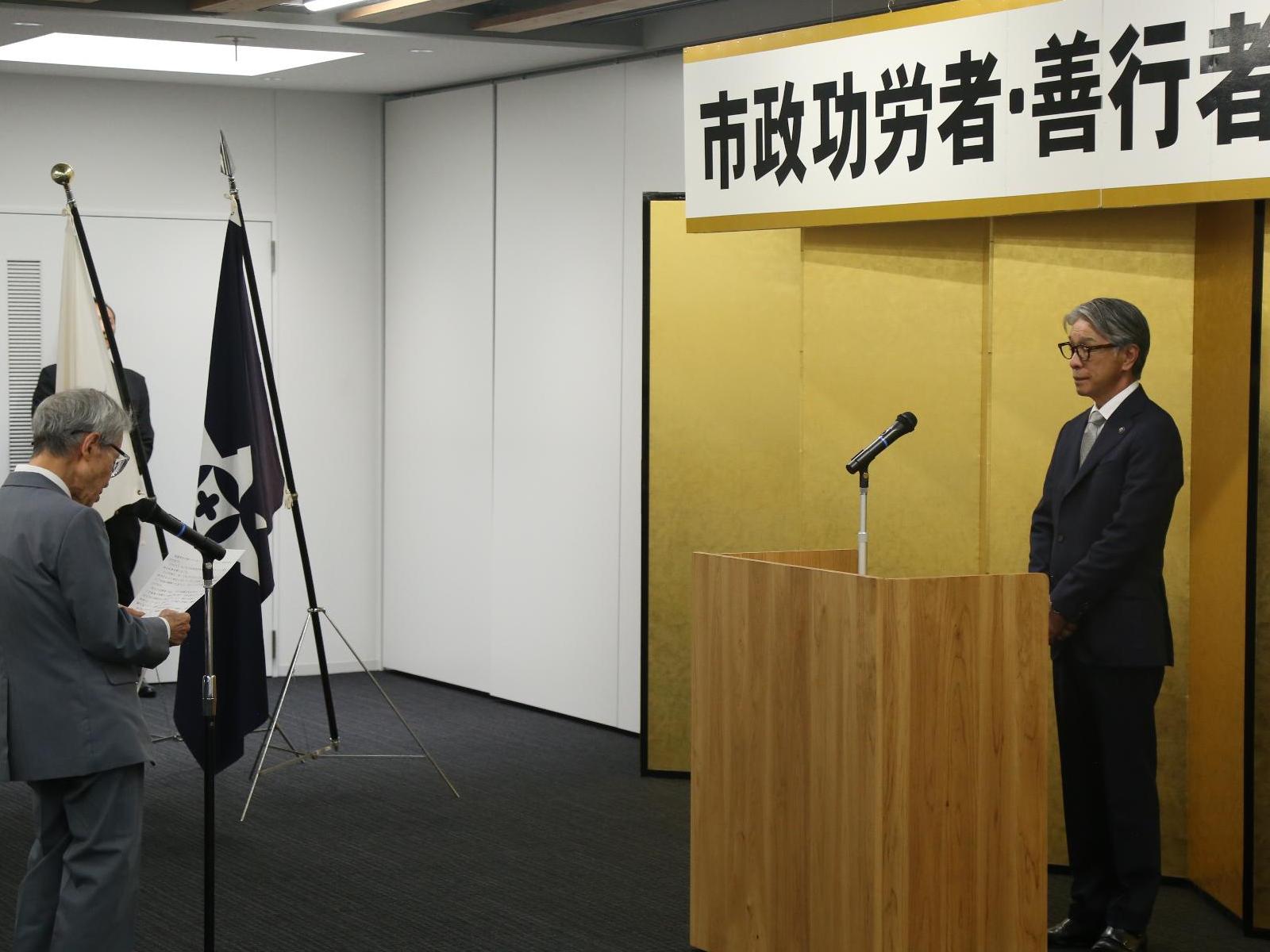 スーツ姿の男性2人が演台を挟んで向かい合い、左の男性がマイクを使って右の男性に話している写真