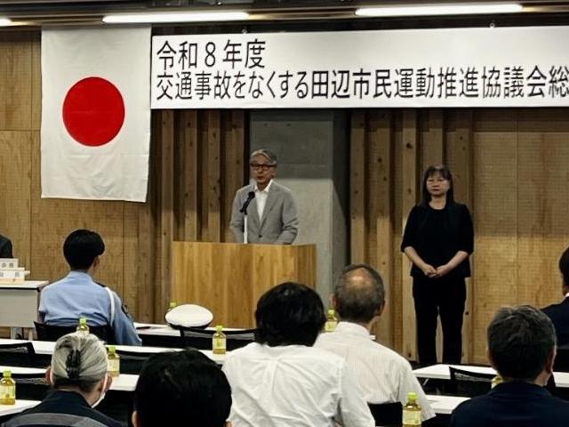 スーツ姿の男性がマイクの前に立ち、出席者に向けて話をしている写真