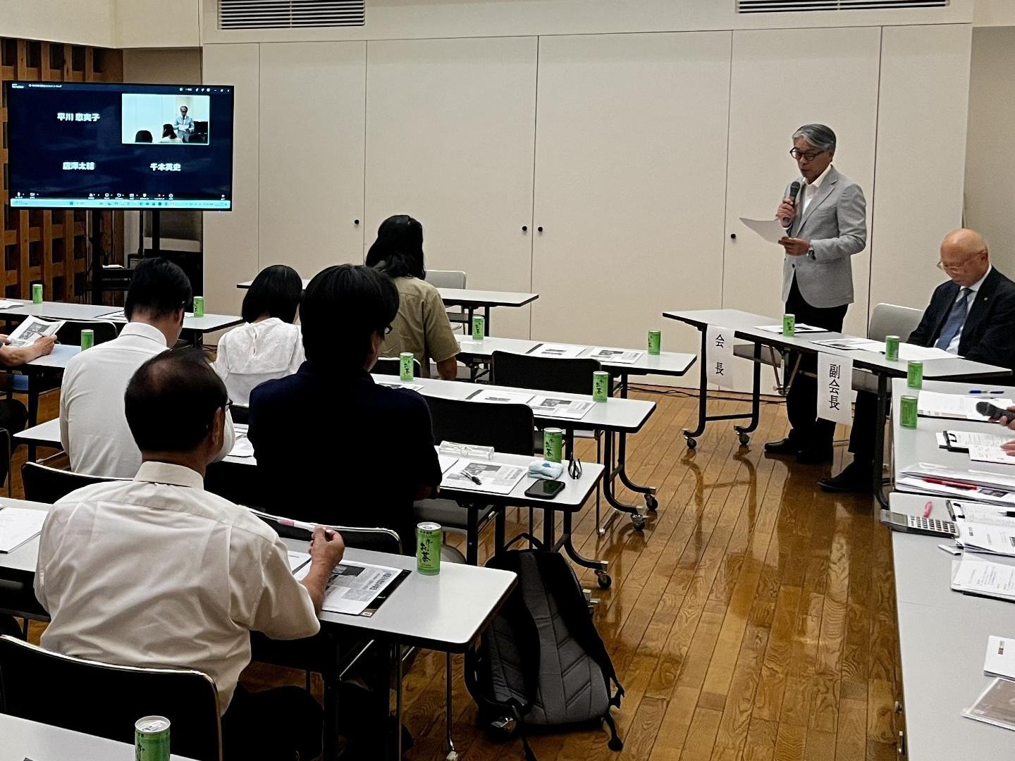 会議室で、前方に立っているスーツ姿の男性の話を出席者が着席して聞いて入る写真