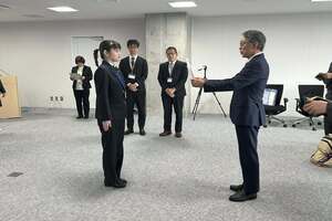 制服姿の女性とスーツ姿の男性が並んでたち、委嘱状らしきものを手渡されている写真