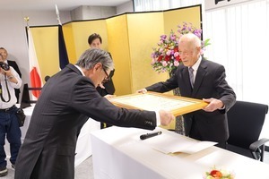 会議室で前方の机にスーツ姿の男性たちが並んで座り、そのうちの一人が資料を説明している写真