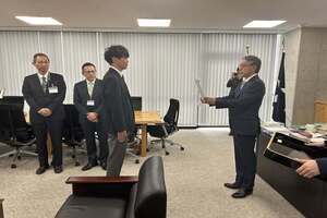 スーツ姿の男性が、前方に立つ人物から書類を両手で受け取っている写真