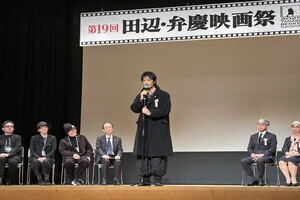 スーツ姿の男性がステージに立って式辞を述べており、客席には多くの参列者が座って参加している写真