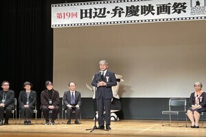 ステージ上にて、吹奏楽団が楽器を構えて記念演奏を行っている様子を捉えた写真