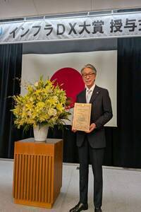 スーツ姿の男性が机の前に座り右手にマイクを持って話をしている写真